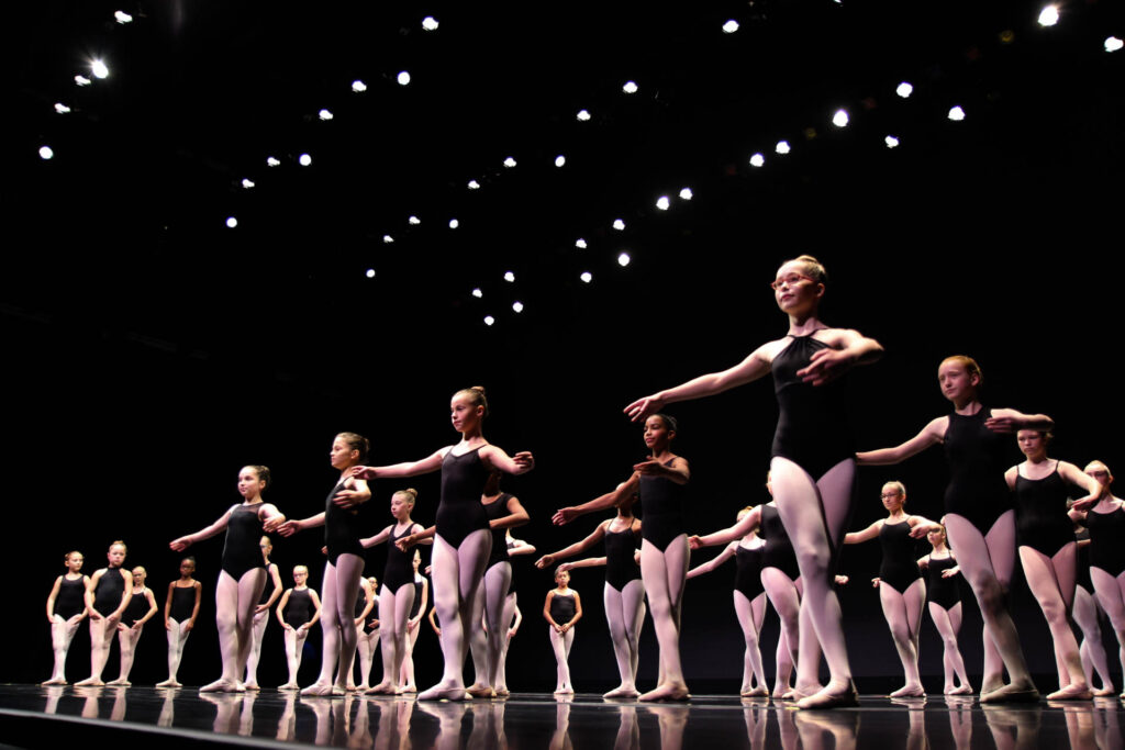 A group of ballet dancers in black leotards and white tights perform a routine on a stage under bright lights.
