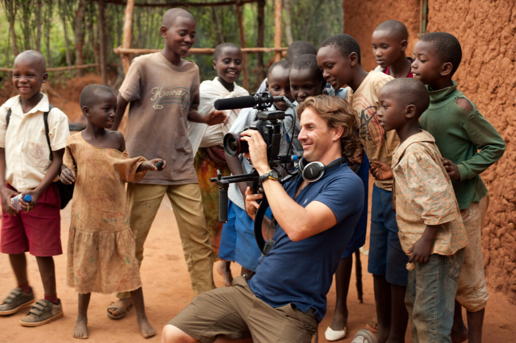A person films with a camera, surrounded by smiling children in a rural outdoor setting.